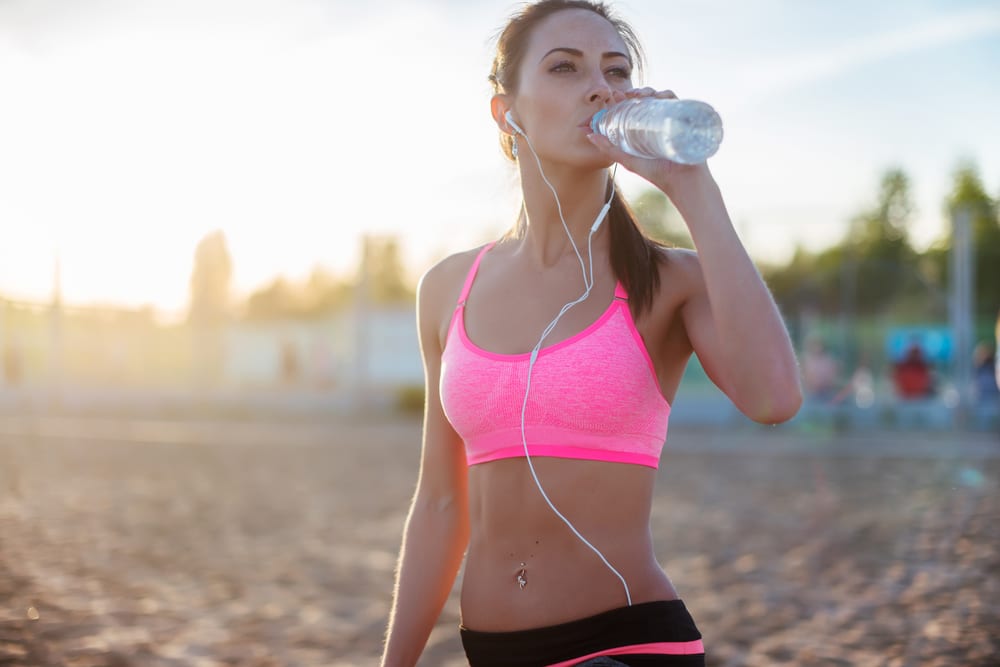 Woman on beach in pink sports bra drinks out of a water bottle.